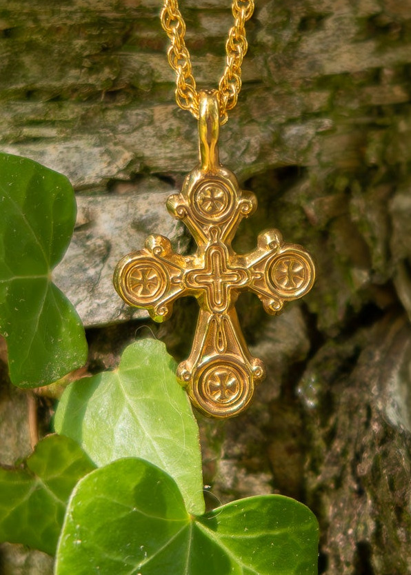 A gold cross pendant necklace with an ornate chain, hanging against a textured tree trunk with green ivy leaves in the foreground.