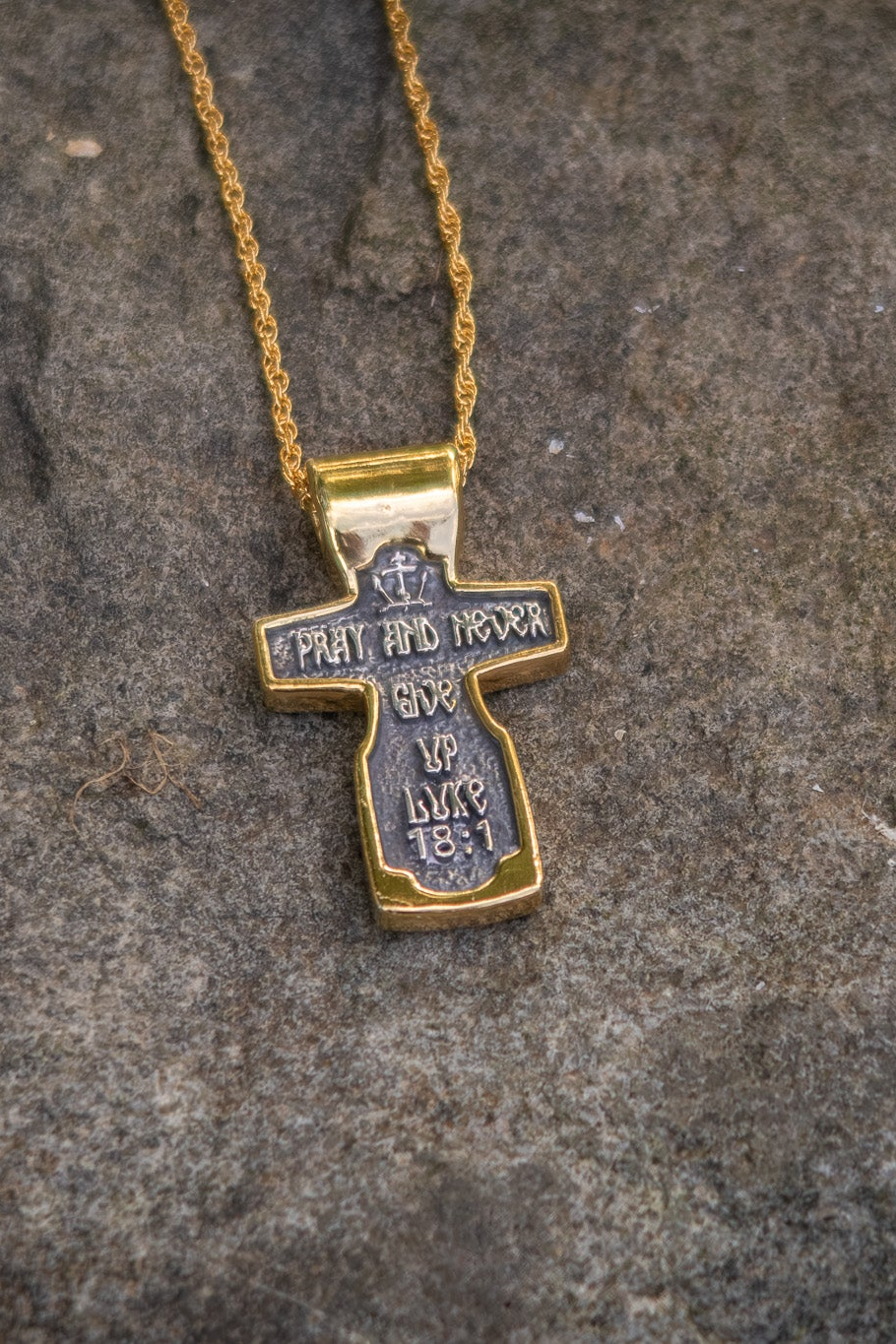 Gold cross pendant with engraved text on a textured stone surface