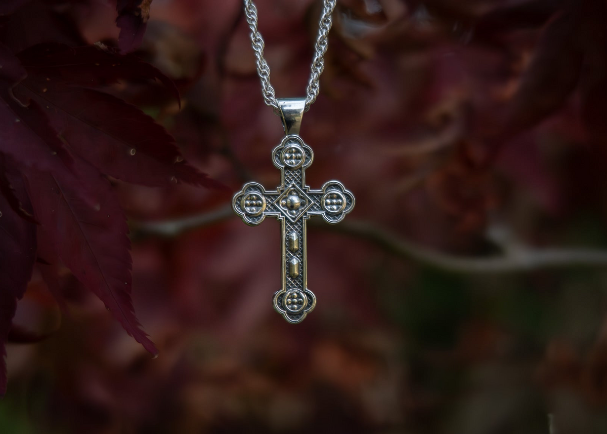A silver-toned ornate cross pendant hanging from a matching chain, set against a blurred background of dark red and brown leaves.