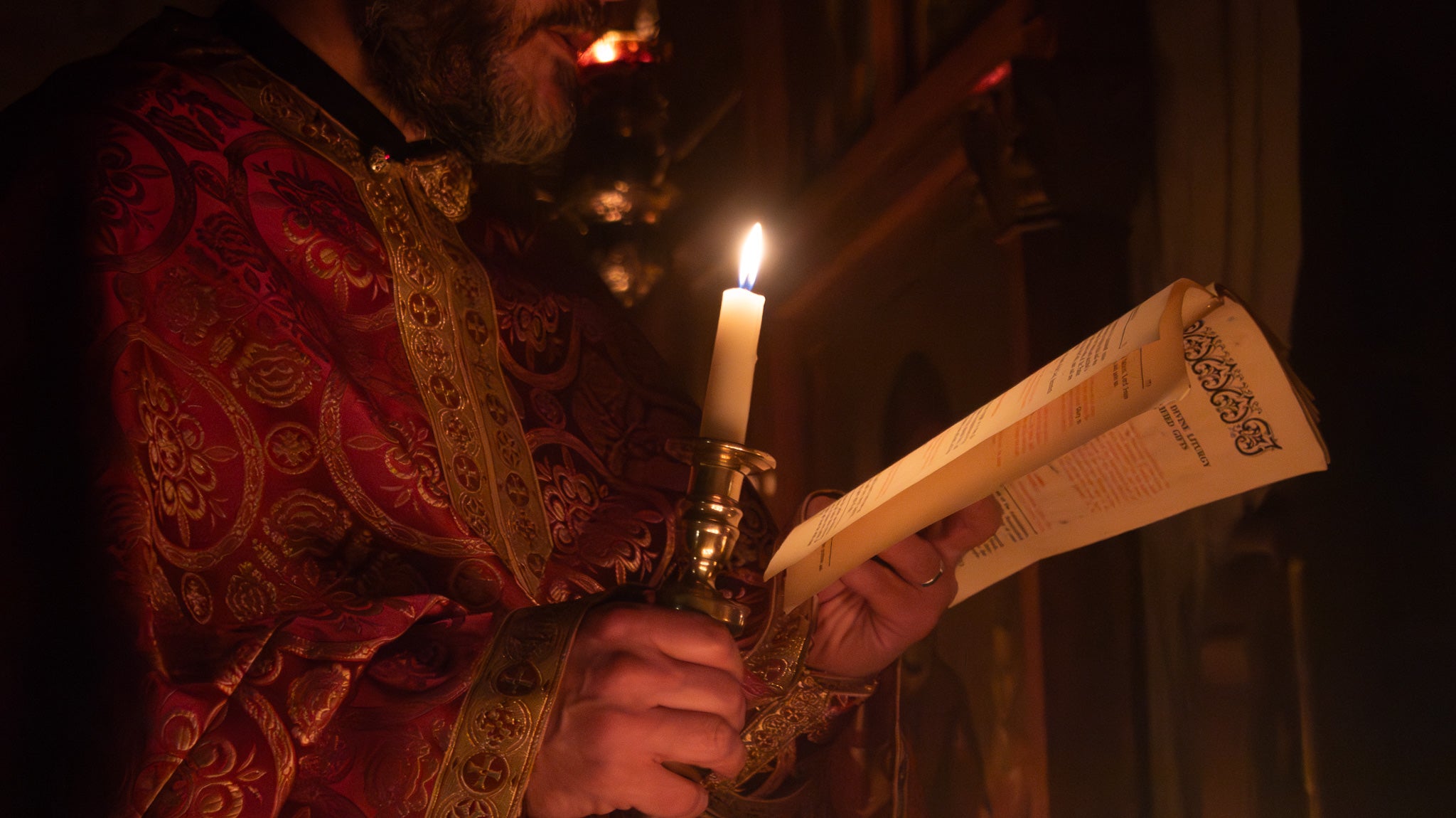 A close-up of a priest in ornate red and gold religious vestments holding a lit candle and an open book in a dimly lit church