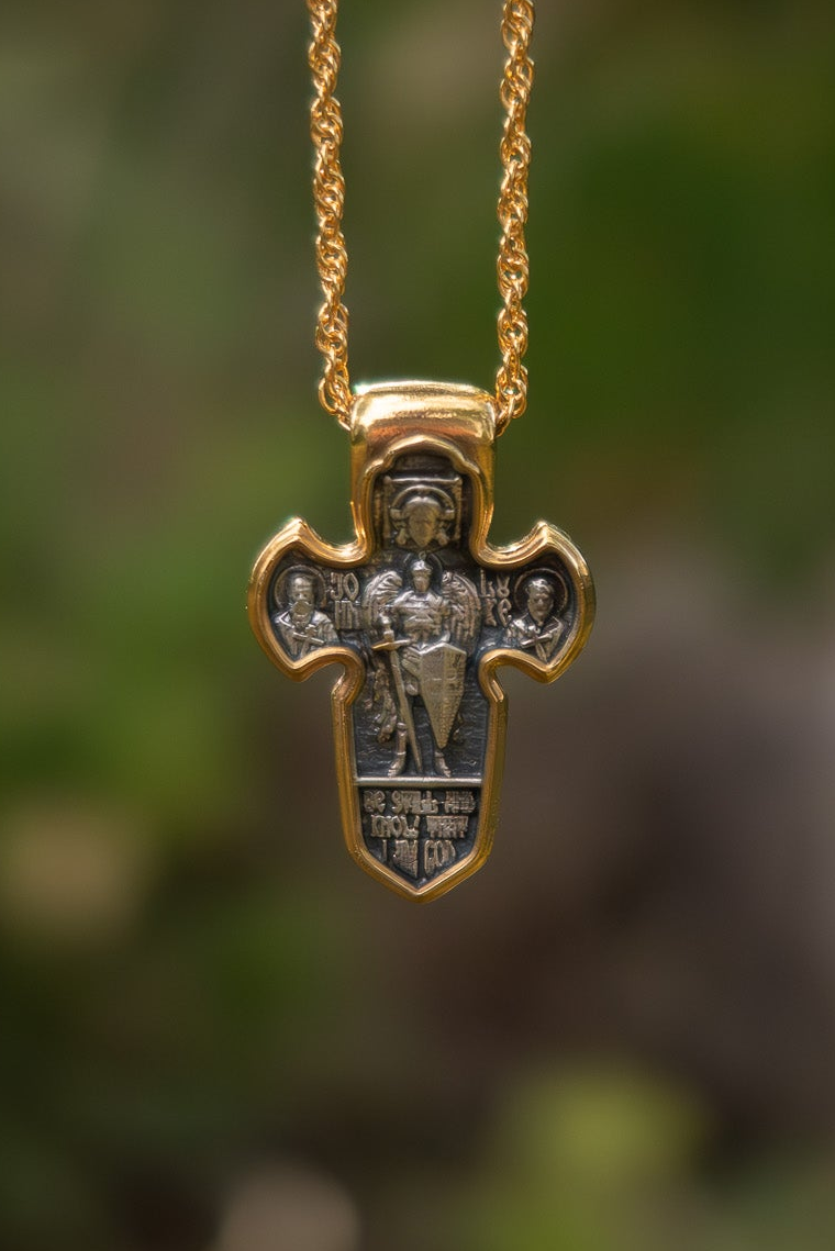 A gold and silver cross pendant necklace featuring a detailed religious icon of Saint George, suspended from a gold chain against a blurred green outdoor background.