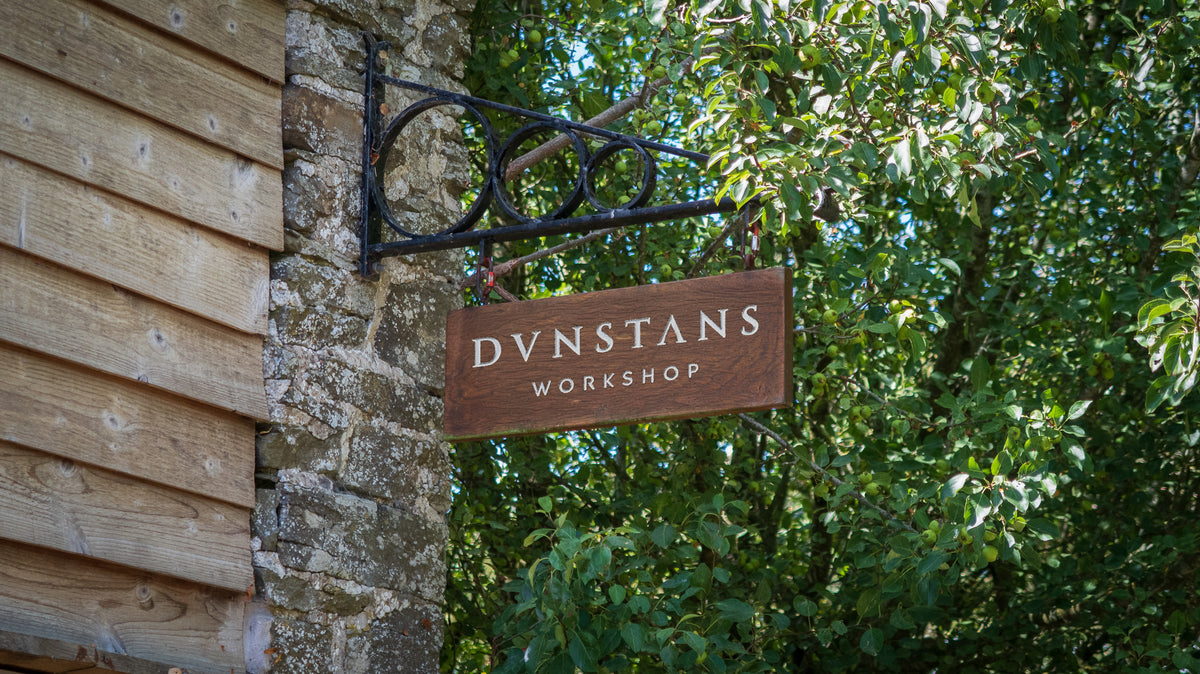 A wooden sign reading 'DUNSTANS WORKSHOP' hangs from a black metal bracket attached to a stone wall beside weathered wood siding, with dense green foliage in the background.
